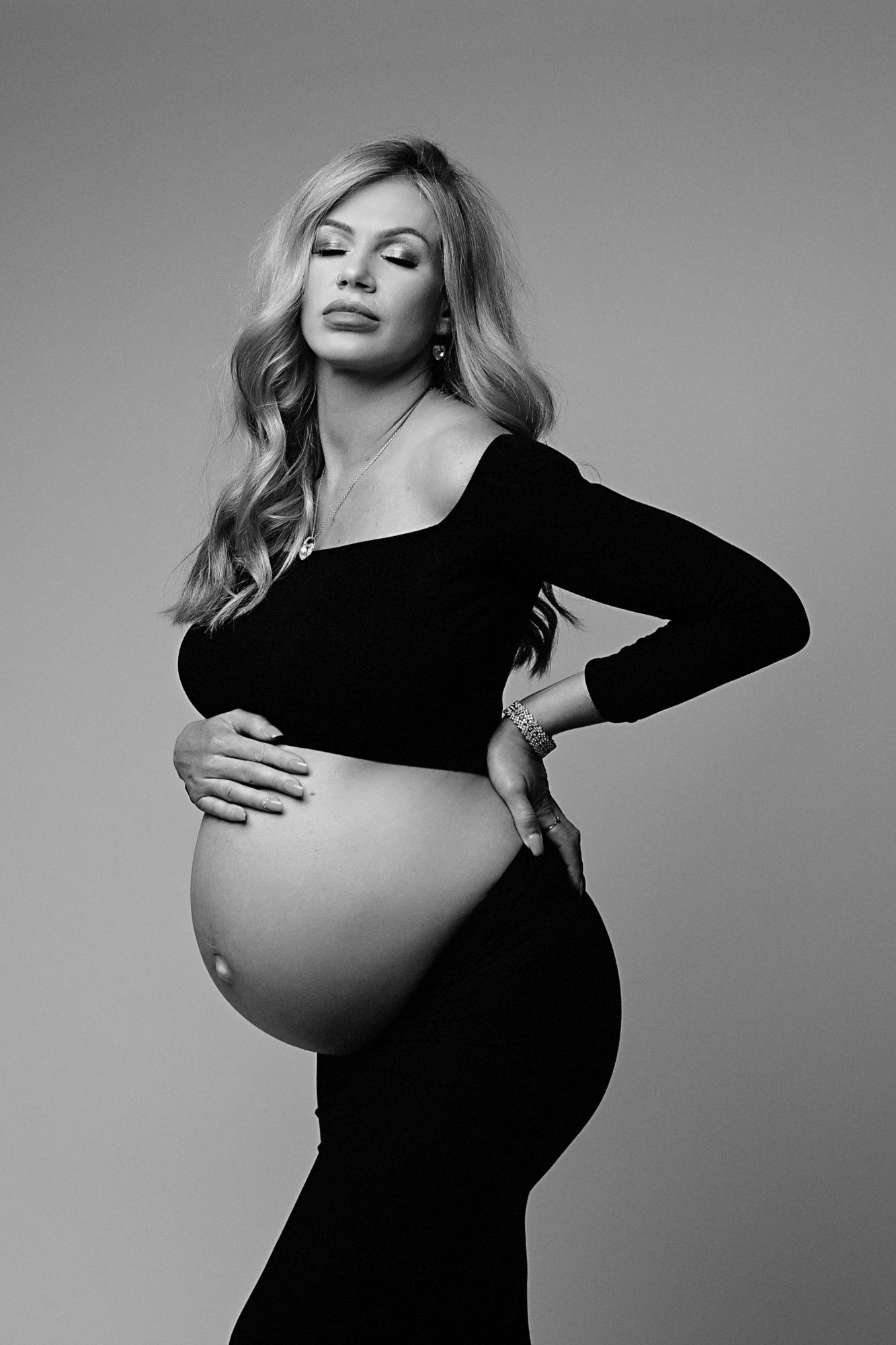 Pregnant Woman Posing with her hand resting gently on her baby bump. She has a black crop top on and skirt. Her hair is blonde and curly.